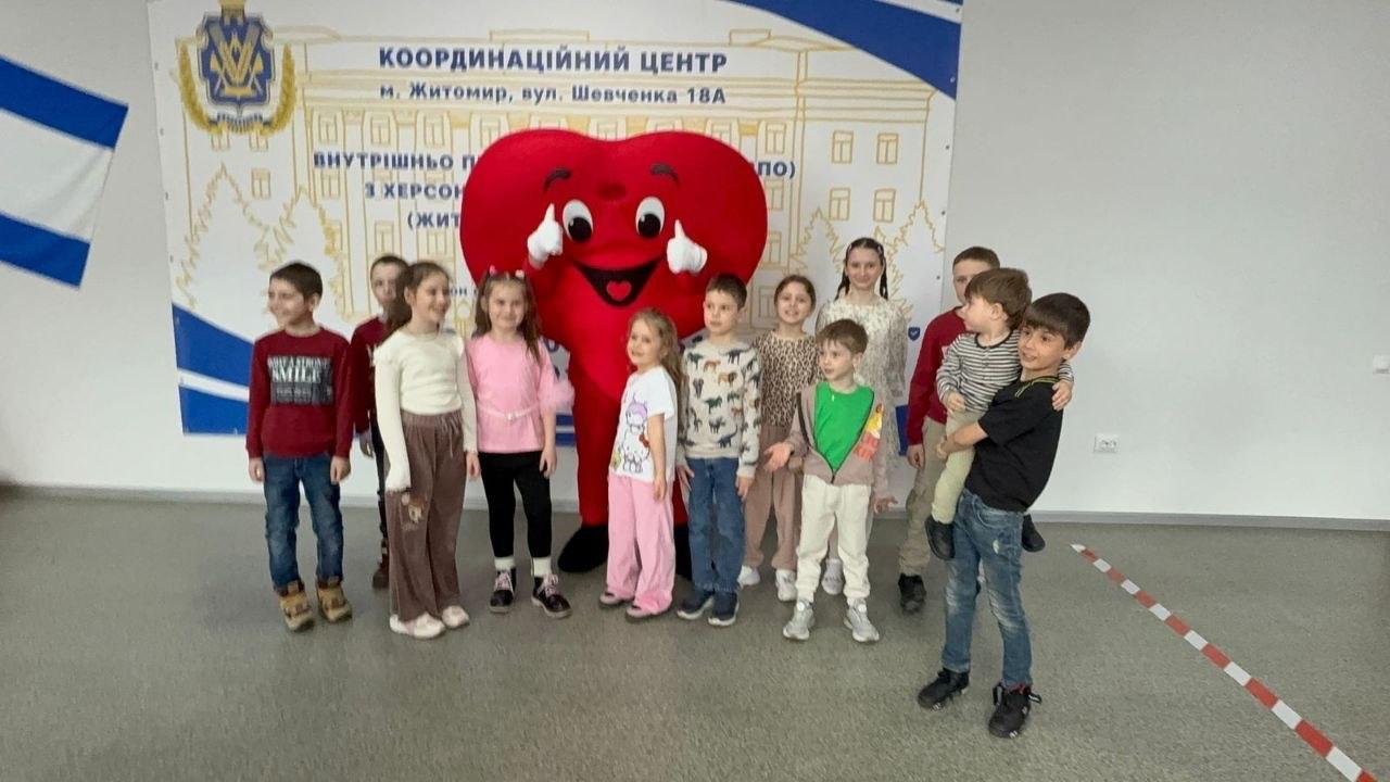 Група дітей позує разом із аніматором у костюмі великого червоного серця в приміщенні Координаційного центру. На задньому плані розміщено банер із написом «Координаційний центр м. Житомир» та державною символікою.