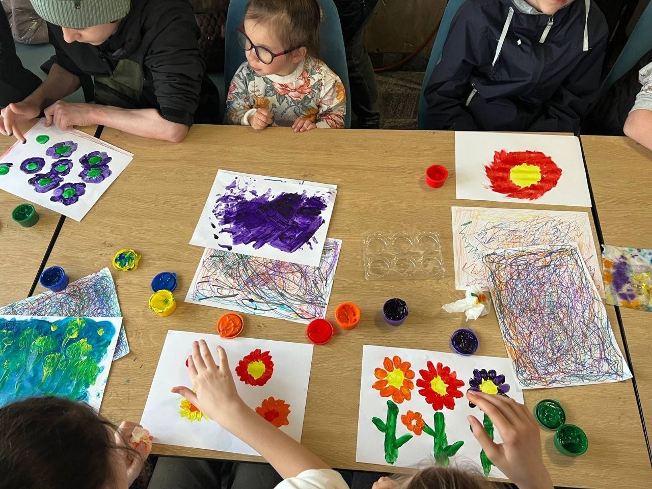 Група дітей та підлітків займається малюванням пальчиковими фарбами за великим дерев'яним столом. На аркушах паперу зображені яскраві квіти, візерунки та абстрактні малюнки насичених кольорів. Поруч лежать відкриті баночки з гуашшю.