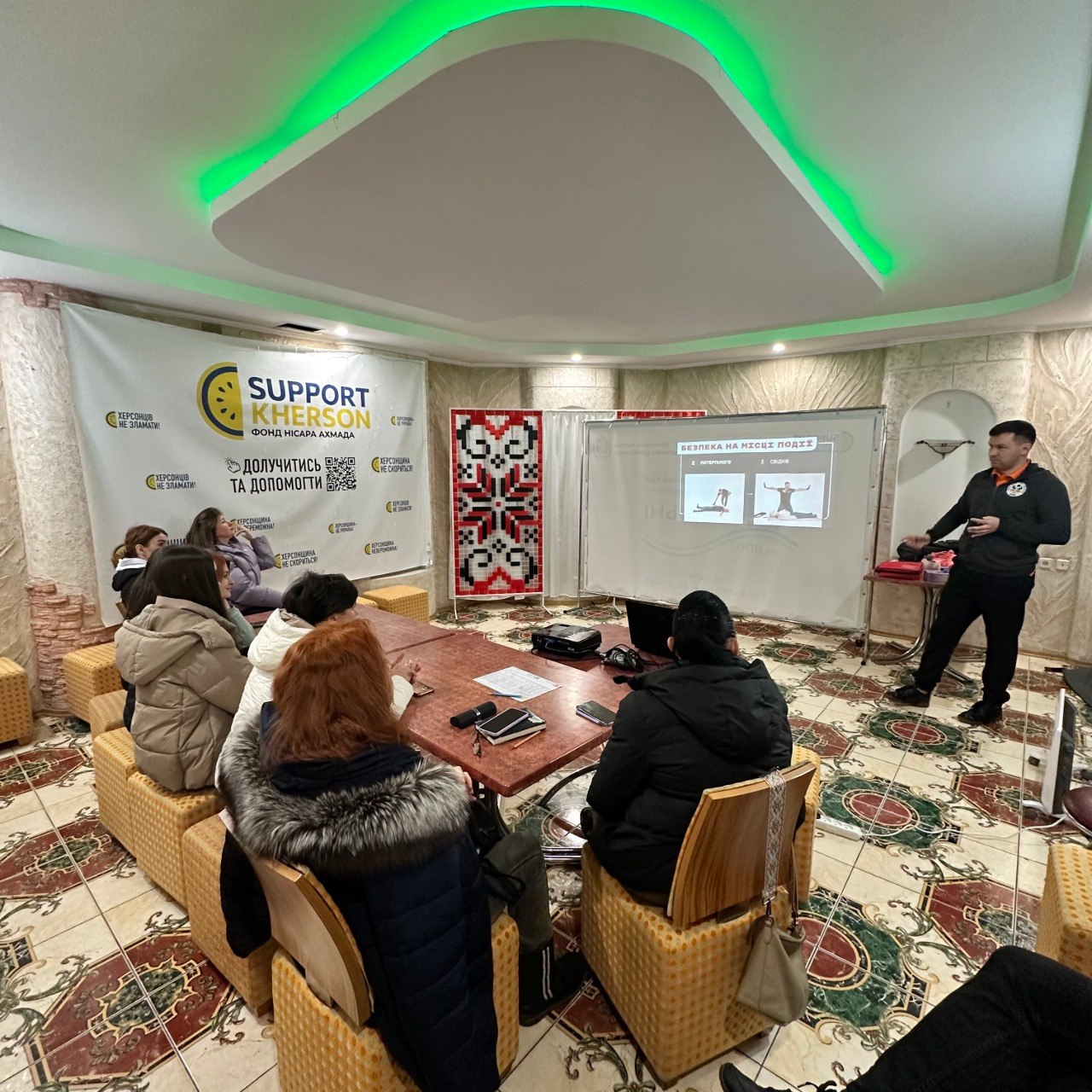 Лекція з безпеки в приміщенні благодійного фонду «SUPPORT KHERSON». Спікер стоїть біля екрана з презентацією про правила поведінки на місці події, а слухачі сидять за столом у верхньому одязі.