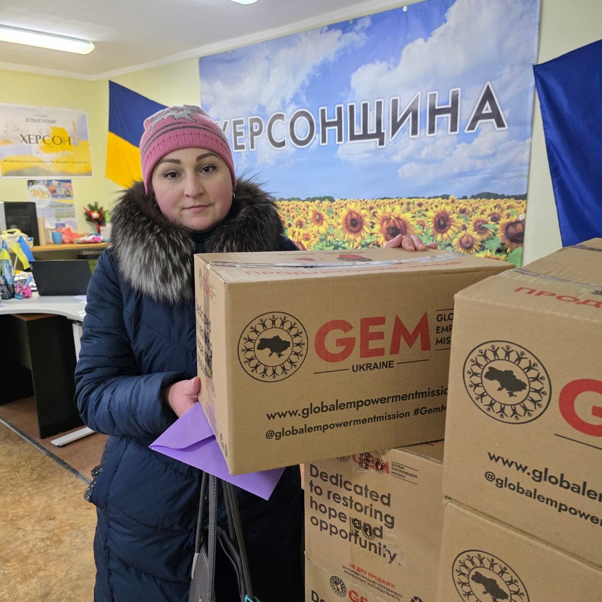 Жінка у зимовому одязі тримає велику коробку з гуманітарною допомогою від організацій GEM та The Howard G. Buffett Foundation. Вона стоїть у приміщенні на фоні банера з написом «Херсонщина» та зображенням соняшників.