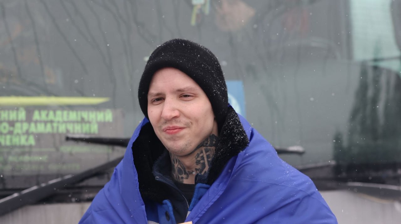 Портрет усміхненого чоловіка в чорній зимовій шапці та синій накидці на плечах. На його шиї видно татуювання, а на задньому плані — вікно автобуса під час снігопаду.