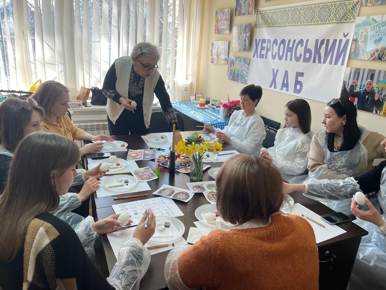 Майстер-клас із розпису писанок у «Херсонському хабі»: група жінок сидить за столом, розписуючи яйця за допомогою писачків та воску під керівництвом майстрині. На столі розкладені зразки орнаментів, фарби та стоїть ваза з жовтими нарцисами.