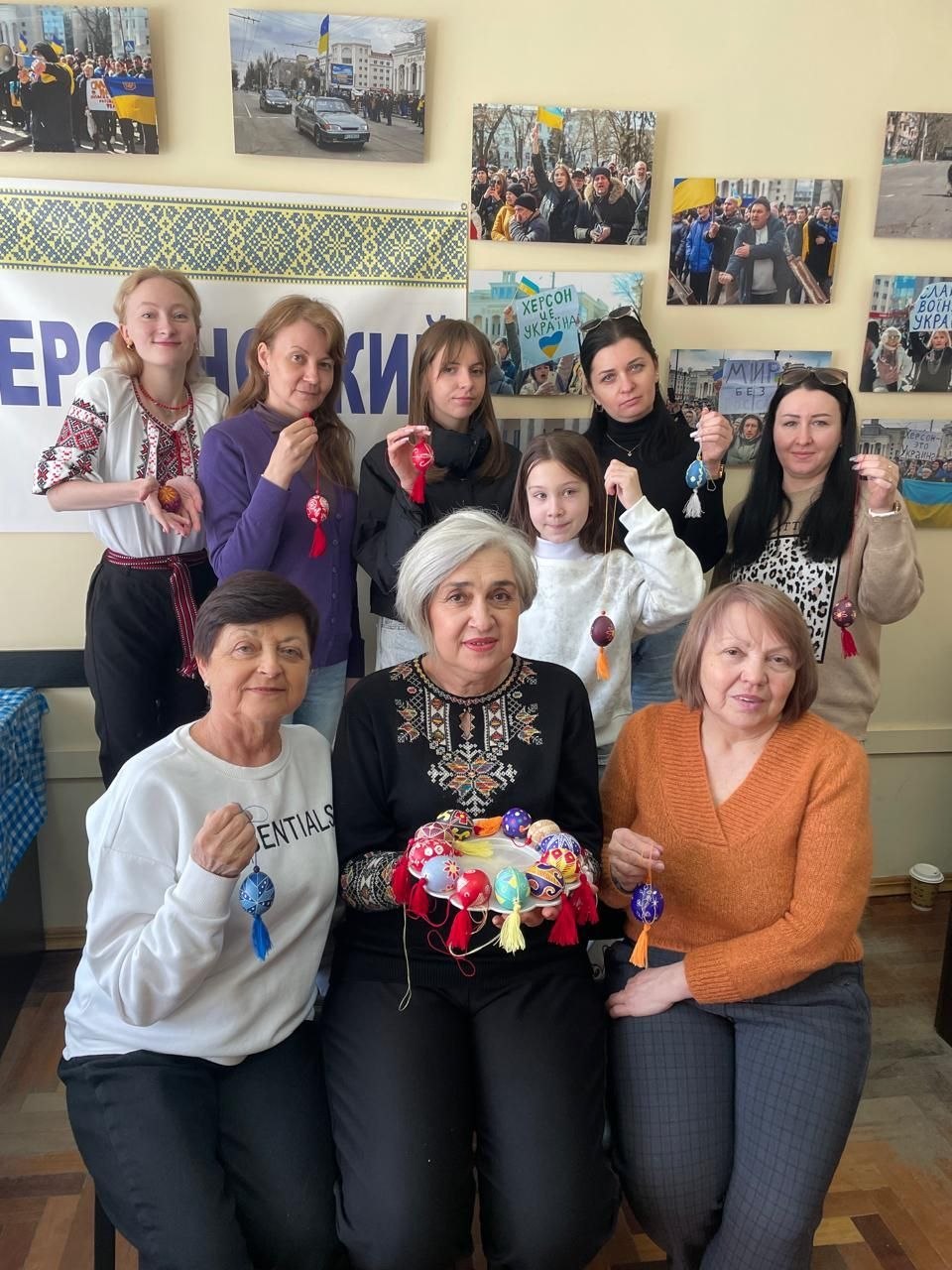 Групове фото учасниць майстер-класу, які позують зі своїми розписаними писанками. На фоні стіни з патріотичними світлинами жінки та дівчата демонструють власноруч створені великодні символи, оздоблені китицями.