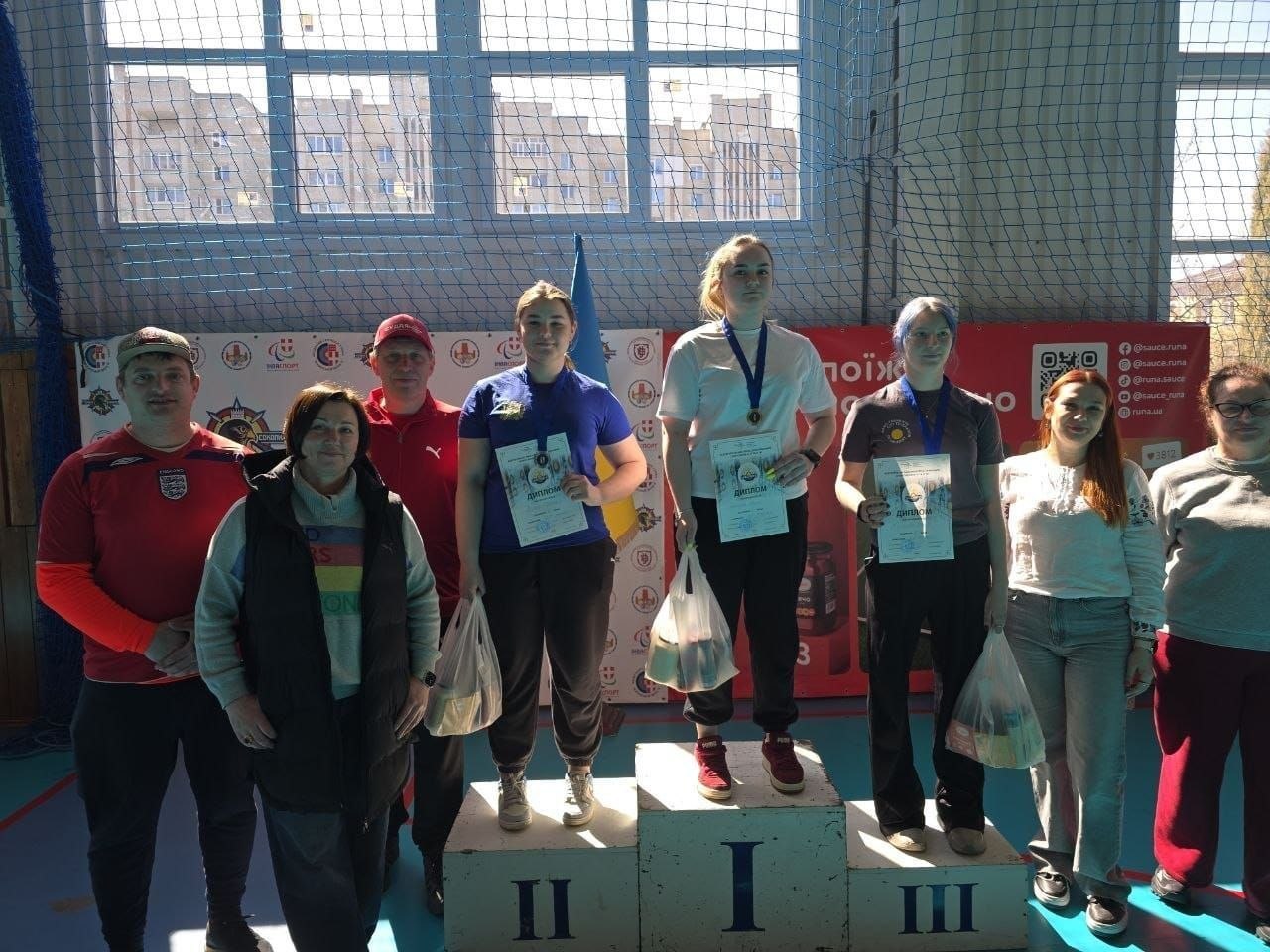 Нагородження призерок спортивних змагань у залі. Три дівчини стоять на п'єдесталі з медалями, дипломами та подарунками, поруч — організатори та інші учасники на фоні банерів і державного прапора.