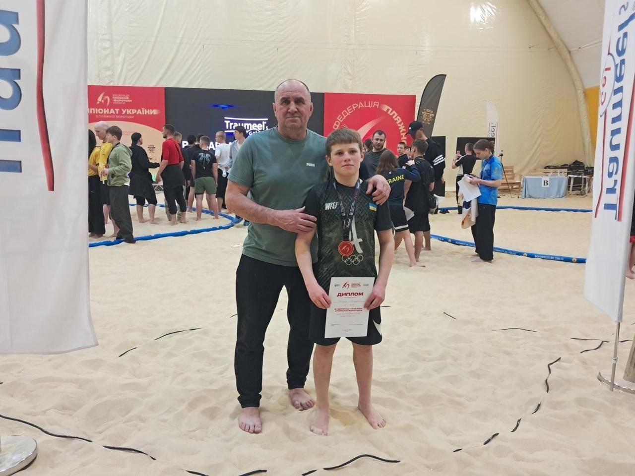 Чоловік та юний спортсмен стоять на піщаному майданчику під час спортивних змагань. Хлопець тримає в руках диплом, а на його шиї висить медаль. На фоні видно інших учасників заходу та банери з офіційною символікою.