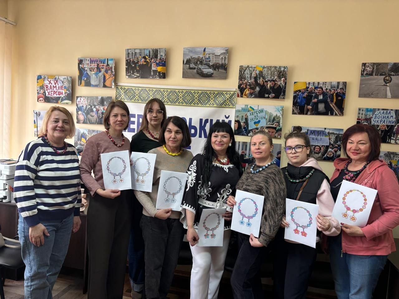 Група жінок стоїть у світлій кімнаті на фоні стіни, прикрашеної численними фотографіями та банером з українським орнаментом. Вони тримають у руках папери з ескізами намист із бісеру (ґерданів), а на деяких учасницях одягнені готові прикраси.