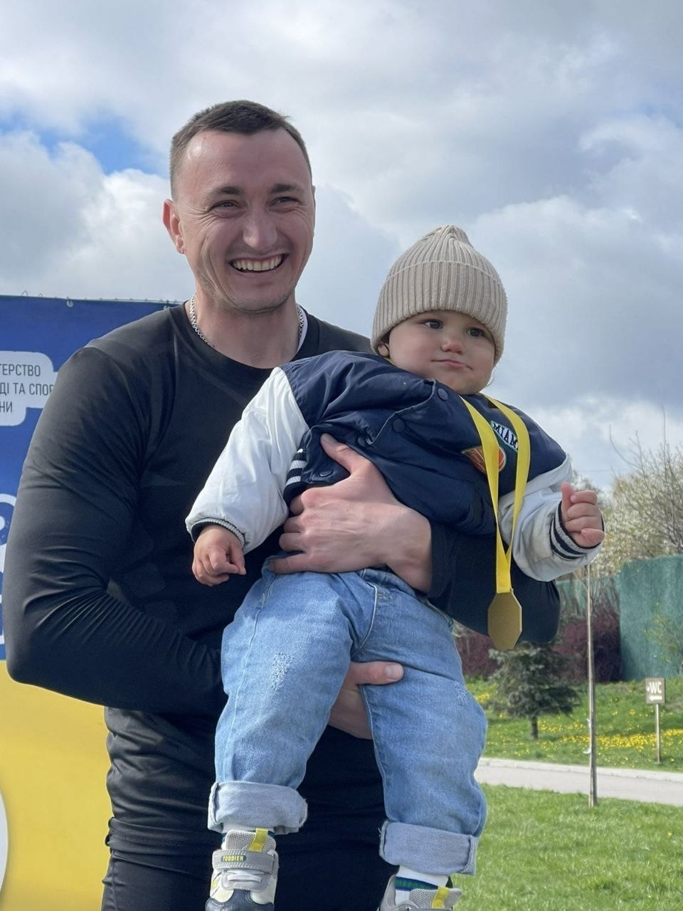 Усміхнений чоловік у чорному спортивному одязі тримає на руках маленьку дитину в куртці та шапці. На шиї дитини висить золота медаль на жовтій стрічці. Вони стоять на вулиці на фоні трави та хмарного неба.