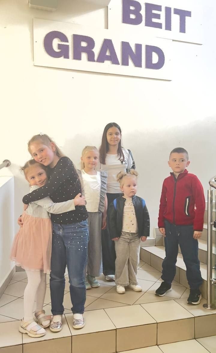 Група дітей різного віку стоїть на сходах у приміщенні на тлі стіни з написами «BEIT» та «GRAND».