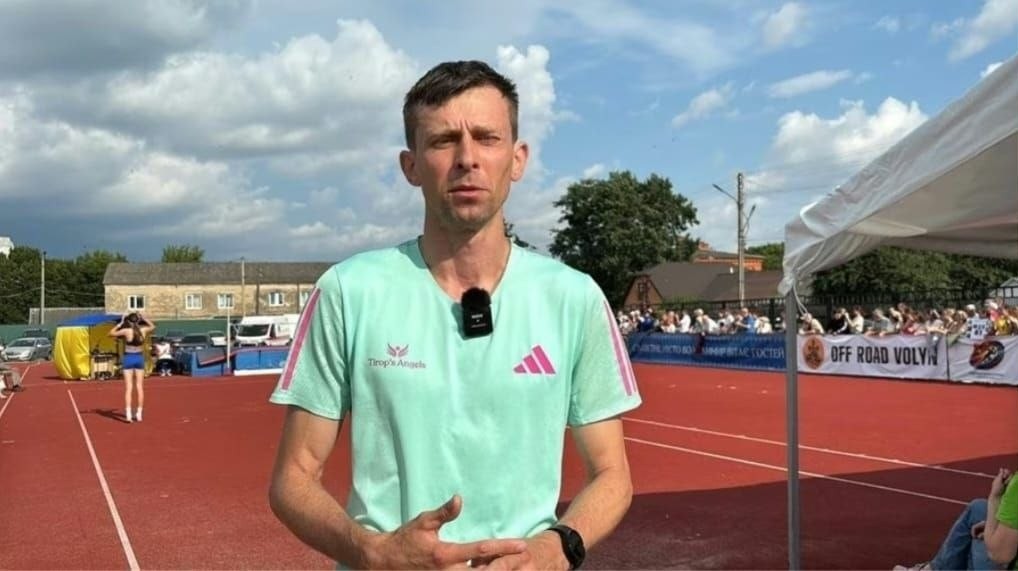 Український легкоатлет, стрибун у висоту Андрій Проценко у світло-зеленій футболці з мікрофоном-петличкою дає інтерв'ю на червоній біговій доріжці стадіону під час змагань.