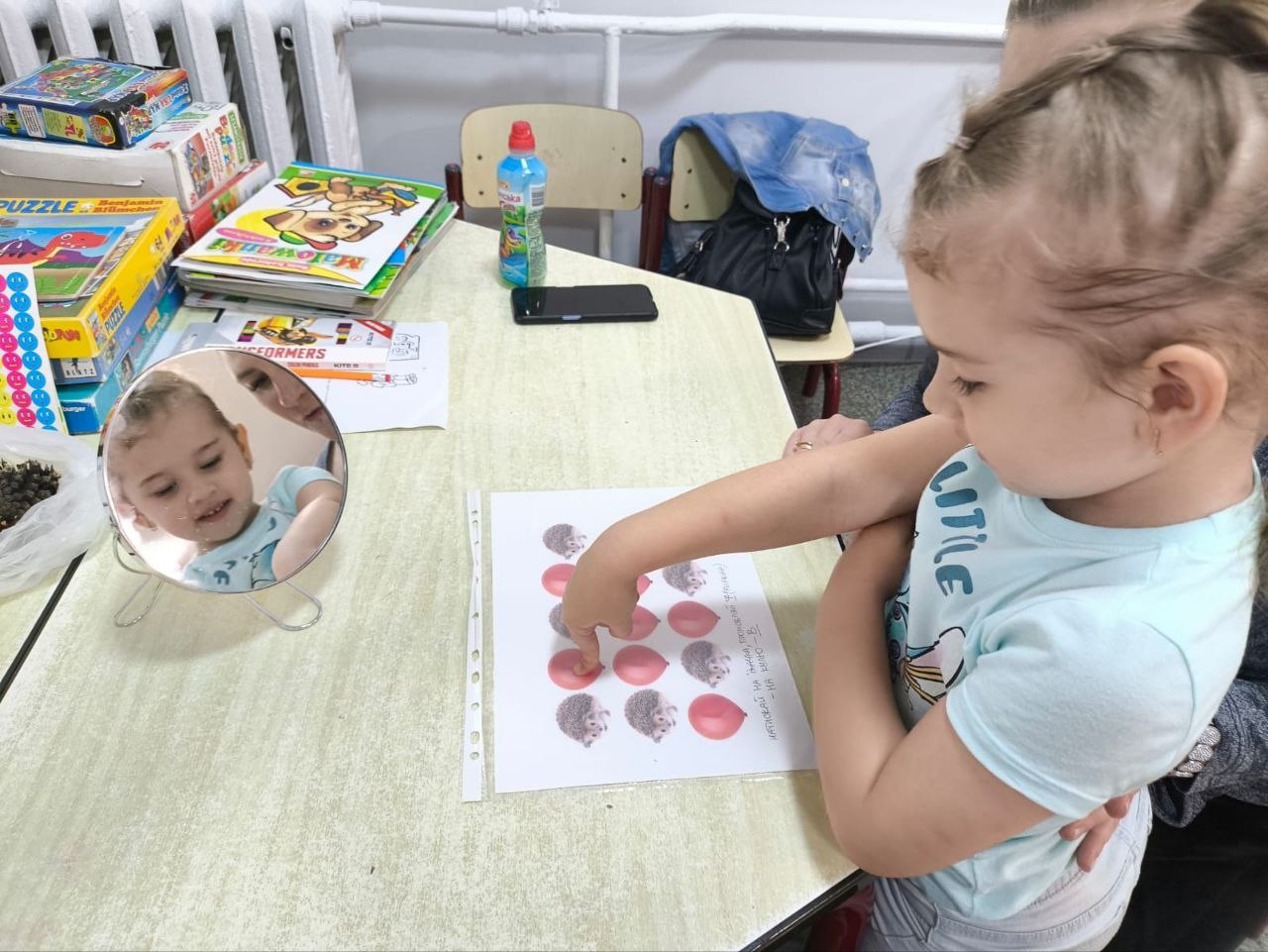 Маленька дівчинка на занятті з логопедом-дефектологом у координаційному центрі «Вільні разом» у Житомирі виконує завдання на папері, показуючи пальчиком на зображення їжачків та кульок. На столі перед нею стоїть кругле логопедичне дзеркало, у якому видно її обличчя, а поруч лежать дитячі розвивальні книги та настільні ігри.