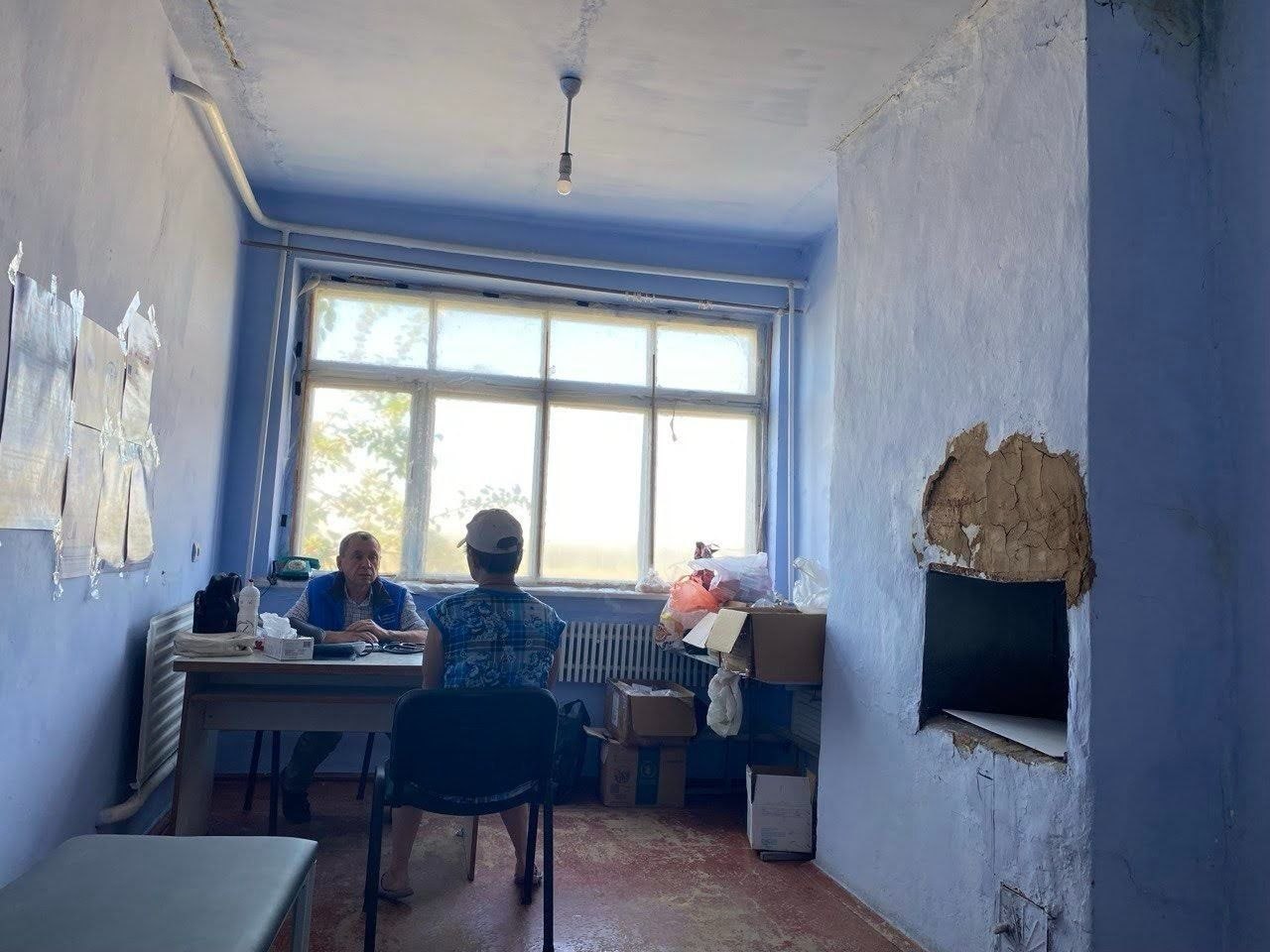 Лікар мобільної медичної команди АСМУ у синьому жилеті консультує пацієнта в облаштованому під кабінет приміщенні з блакитними стінами в одному з прифронтових сіл Херсонщини.