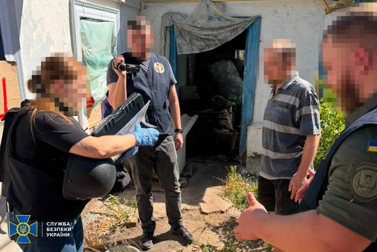 Троє співробітників СБУ та контррозвідки проводять обшук та процесуальні дії на подвір'ї приватного будинку у Херсоні в присутності підозрюваного чоловіка у смугастій футболці. Співробітниця ліворуч у блакитних медичних рукавичках тримає захисний шолом та спеціальний кейс, оперативник по центру знімає процес на портативну відеокамеру, а правоохоронець праворуч спілкується з фігурантом. Обличчя всіх чотирьох людей розмиті з метою збереження таємниці слідства, у лівому нижньому кутку розміщено офіційний логотип Служби безпеки України.