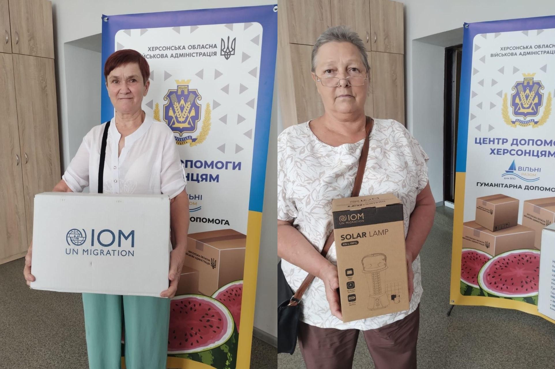 Колаж із двох фотографій, що ілюструють видачу гуманітарної допомоги для внутрішньо переміщених осіб із Херсонської області в координаційному центрі Вільні разом. На лівому знімку жінка старшого віку стоїть у приміщенні хабу та тримає велику білу коробку з логотипом Міжнародної організації з міграції IOM UN Migration, у якій міститься зимовий постільний набір. На правому знімку інша жінка похилого віку тримає картонну коробку від тієї ж організації з написом Solar Lamp, призначену для отримання світлодіодної лампи на сонячній батареї. Обидві жінки позують на тлі вертикальних інформаційних банерів Херсонської обласної військової адміністрації Центр допомоги херсонцям Вільні разом. Гуманітарна допомога. Видача цих наборів та ліхтарів спрямована на матеріальну підтримку переселенців, забезпечення їхніх базових побутових потреб в енергонезалежному освітленні та підвищення життєстійкості громадян під час війни.