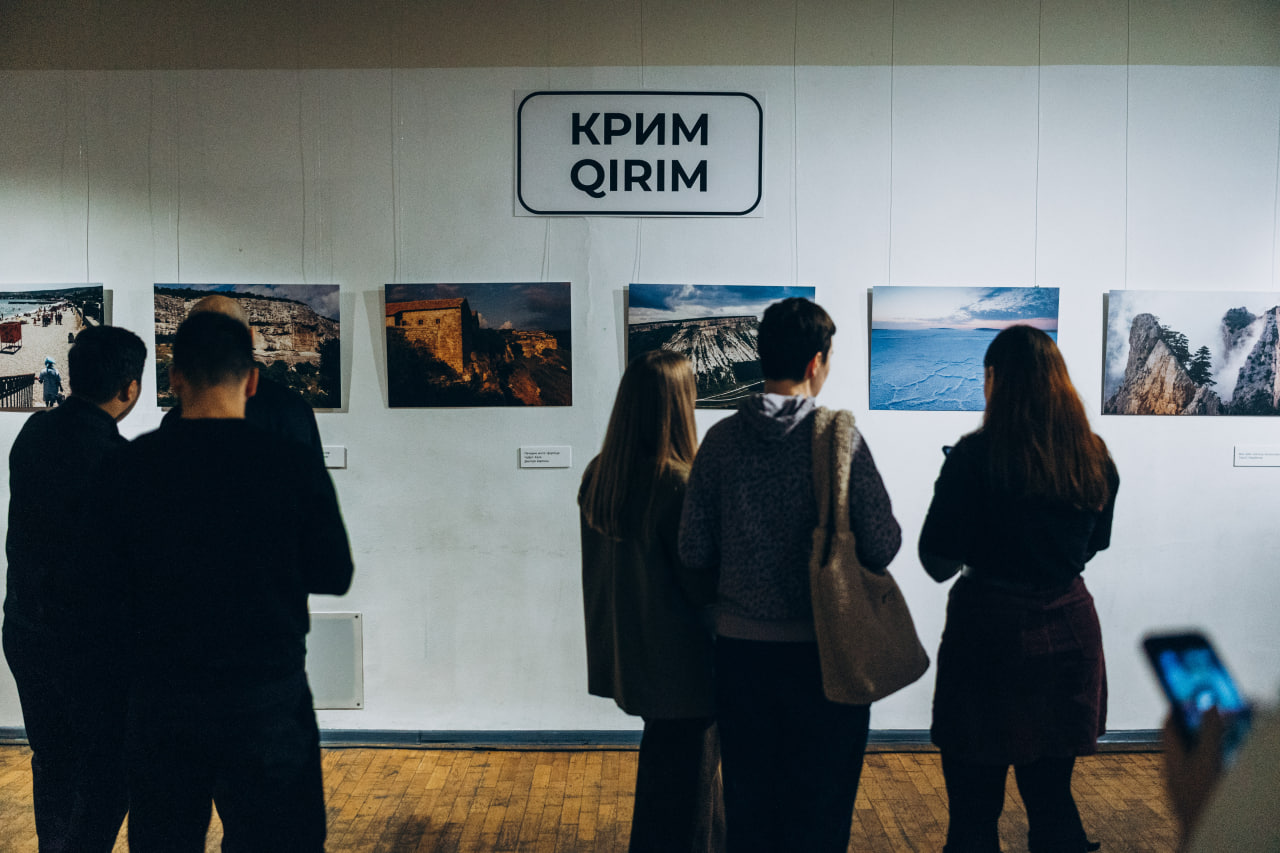 Відвідувачі виставки розглядають експозицію фотографій із краєвидами Криму під вказівником «КРИМ / QIRIM». На стіні розміщено шість світлин, що демонструють природу та архітектуру півострова.