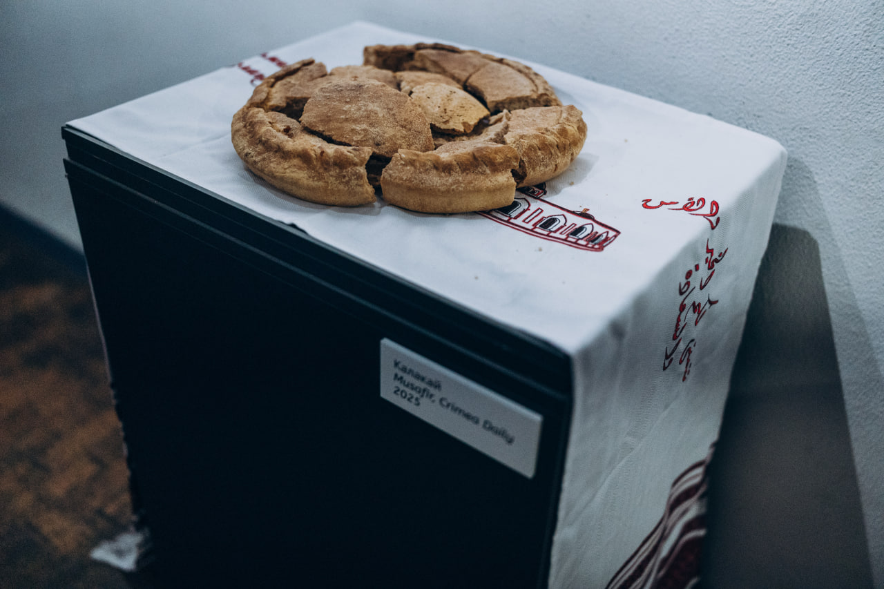 Традиційний кримськотатарський хліб калакай, розламаний на частини, лежить на вишитому рушнику поверх музейної тумби. Поруч розміщено етикетку з назвою експоната та роком створення — 2025.