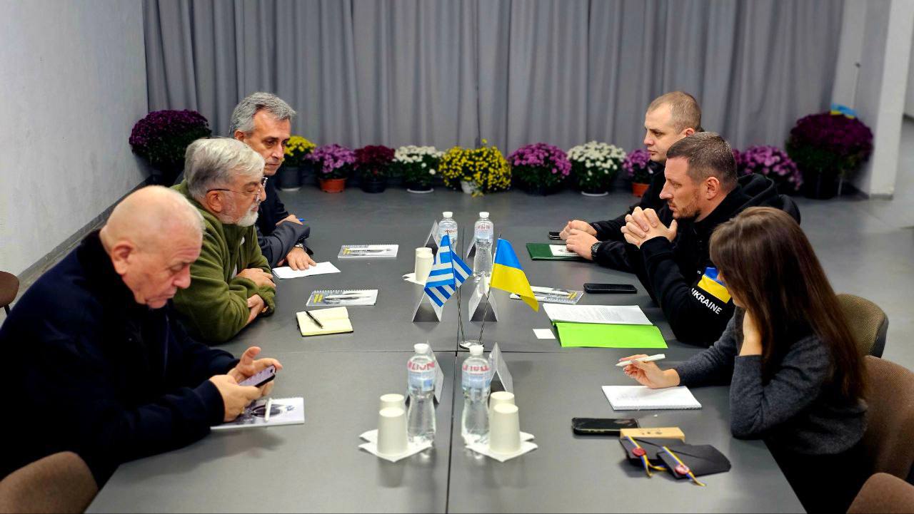 Зображення: Зустріч за столом, де присутні сім осіб. На столі розміщені прапори Греції та України, пляшки з водою та папери. Двоє чоловіків ліворуч у профіль, один з них дивиться на свій телефон. Четверо людей справа сидять обличчям до камери. На задньому плані — квіткові композиції.