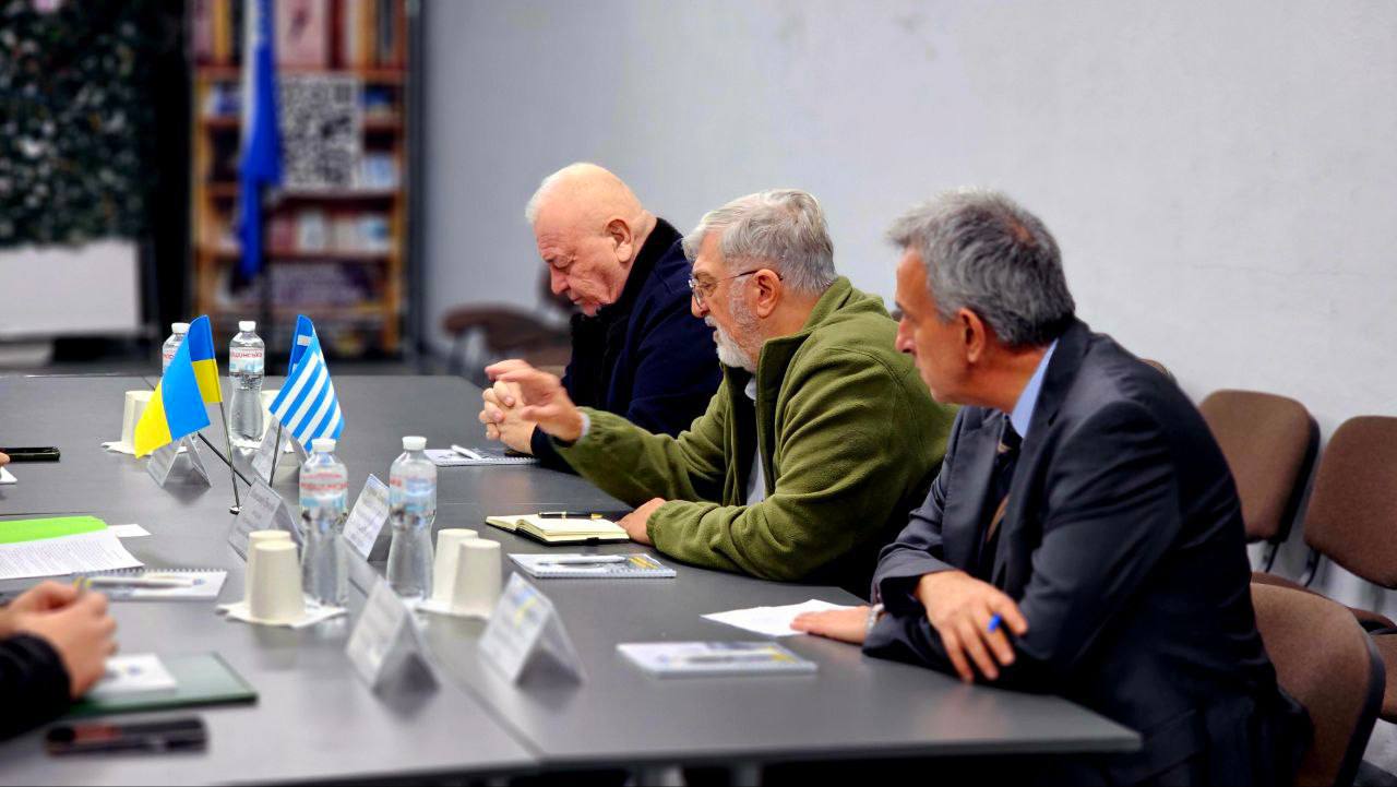 Троє старших чоловіків сидять за столом переговорів. Чоловік у центрі, одягнений у зелену куртку, щось пояснює, жестикулюючи правою рукою. Чоловік ліворуч нахилив голову, а чоловік праворуч у костюмі дивиться вперед. На столі перед ними стоять мініатюрні прапори України та Греції, пляшки з водою та папери.