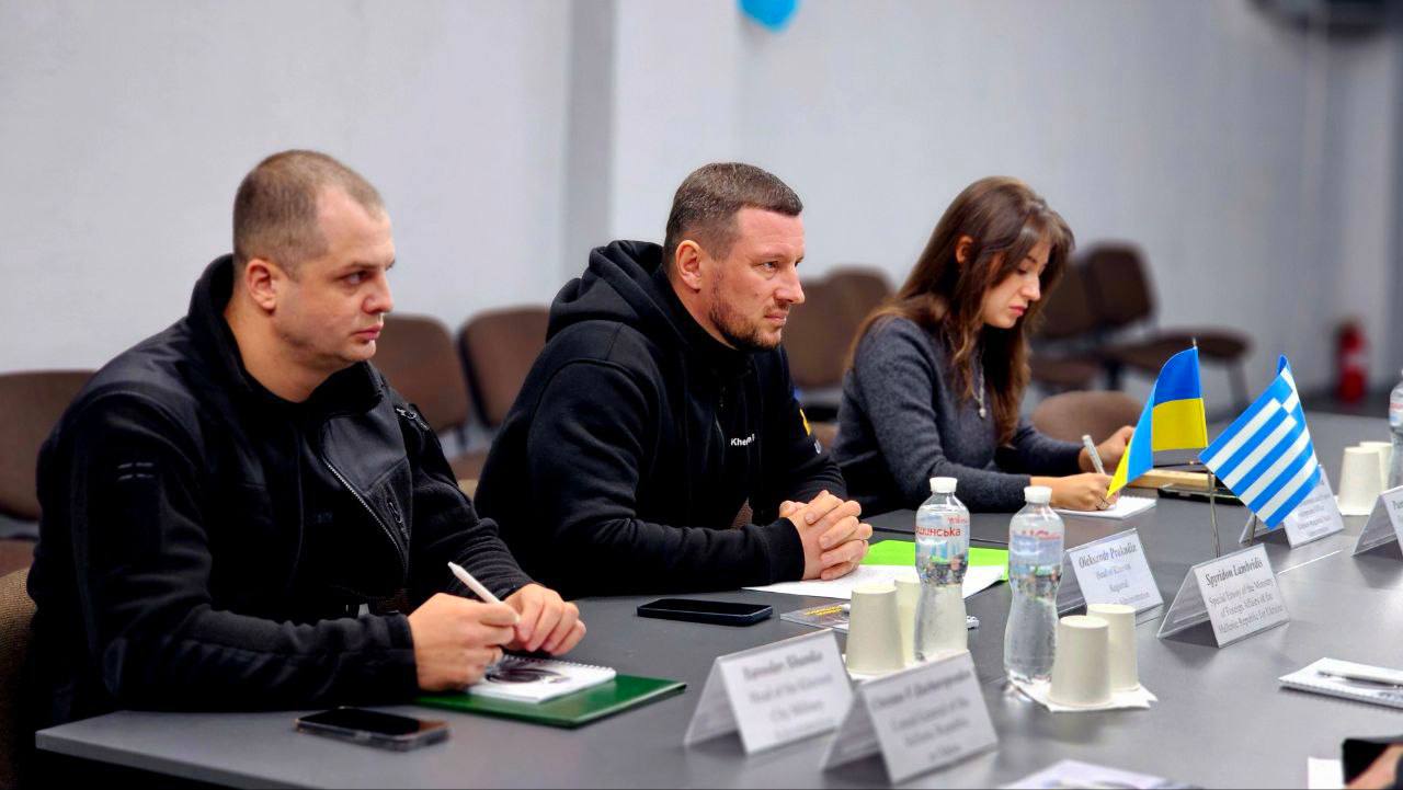 Троє людей сидять за столом під час зустрічі. Усі дивляться вправо. Чоловік ліворуч у чорній куртці, посередині — чоловік у чорному худі, праворуч — жінка, яка щось записує. На столі перед ними стоять мініатюрні прапори України та Греції, а також таблички з іменами та посадами.