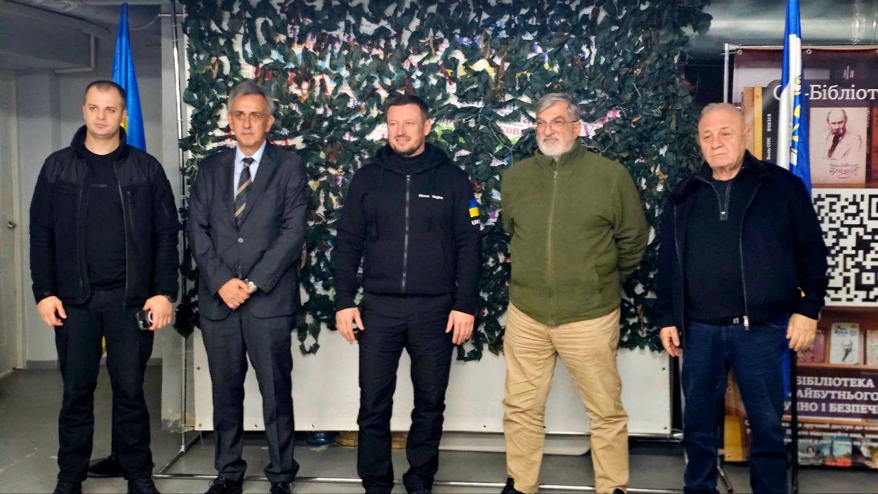 П'ятеро чоловіків стоять у ряд, обличчям до камери. Вони знаходяться в приміщенні на тлі стіни, задрапірованої маскувальною сіткою, а також книжкової полиці з QR-кодом і написом: Бібліотека майбутнього праворуч. Зліва стоїть синьо-жовтий прапор. Чоловіки одягнені у цивільний одяг: двоє — в чорному, один — у костюмі, ще один — у зеленій куртці та бежевих штанях, і один — у чорній кофті та джинсах.