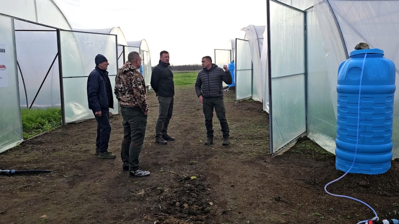 Четверо чоловіків спілкуються біля входу в теплиці поруч із великою синьою бочкою для води.