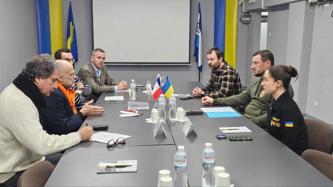 Група з восьми людей (чоловіків і однієї жінки) засідає за великим прямокутним столом, обговорюючи щось, із прапорами України та Франції на столі. На задньому плані видно великий проекційний екран та прапори України.