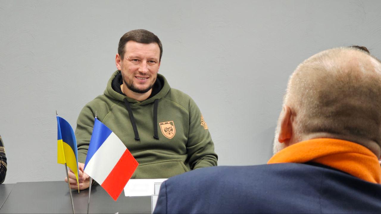 Портретний знімок чоловіка у світло-зеленому худі із шевроном, який сидить за столом і посміхається, дивлячись на співрозмовника, спина якого (з помаранчевим шарфом) знаходиться на передньому плані. На столі видно невеликі прапори України та Франції.