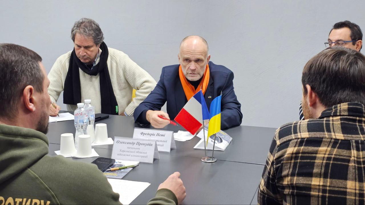 На середньому плані зображено чоловіка у темно-синьому костюмі та помаранчевому шарфі, який сидить за столом, тримаючи ручку і дивлячись прямо на камеру. Перед ним на столі стоять мініатюрні прапори України та Франції. Також на столі видно таблички з іменами.