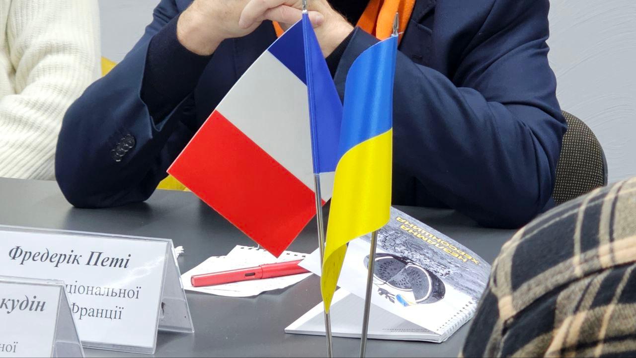 Фокус на мініатюрних прапорах Франції та України, які стоять на столі під час зустрічі. Частково видно таблички з іменами та руки людини у темно-синьому піджаку з помаранчевим елементом одягу.