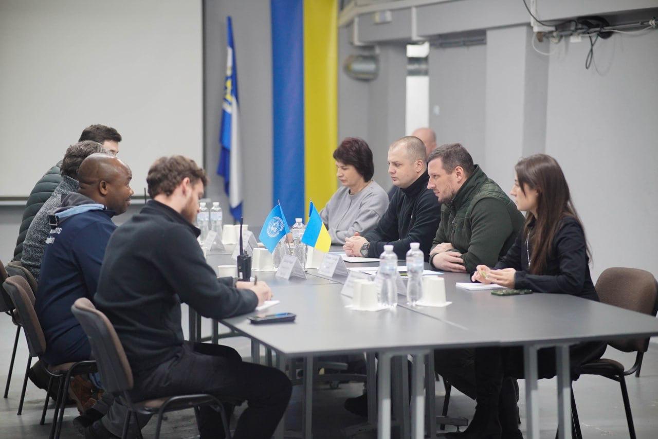 Група людей сидить за довгим столом у приміщенні на нараді. На столі розташовані прапори України та ООН. На задньому плані видно великий український прапор.