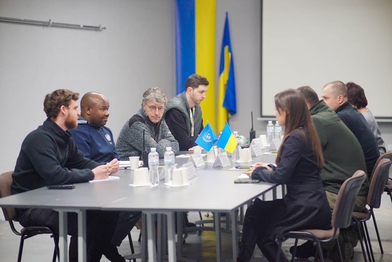 Група з восьми людей (чоловіків і жінок) сидить за столом для нарад. У центрі столу розташовані мініатюрні прапори України та ООН. На задньому плані видно великий український прапор.