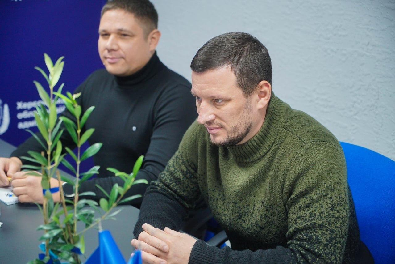 Двоє чоловіків сидять за столом у приміщенні. Чоловік на передньому плані, одягнений у зелений светр, дивиться вниз і тримає руки складеними. Чоловік позаду, одягнений у чорний светр, дивиться вперед. На столі перед ними стоїть невелика рослина з зеленим листям (схожа на оливкове дерево).