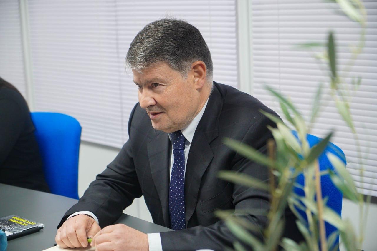 Чоловік у діловому костюмі та краватці сидить за столом для нарад, уважно слухаючи або розмірковуючи. На передньому плані праворуч видно гілку оливкового дерева.