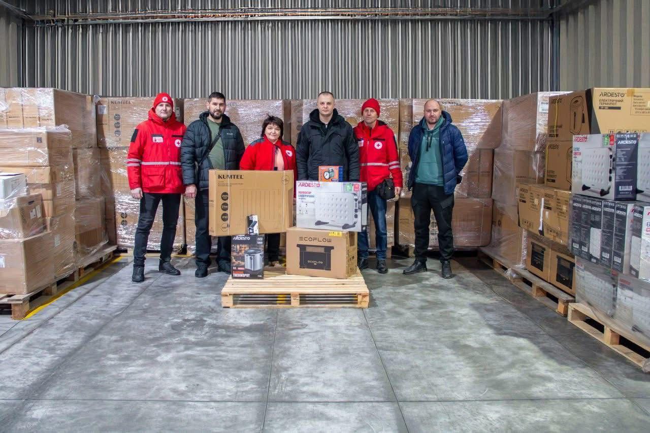 Шестеро людей у теплому верхньому одязі (двоє з них у червоній формі з логотипами Червоного Хреста) стоять на складі на фоні численних коробок із гуманітарною допомогою. Перед ними на дерев’яній палеті виставлені зразки техніки: обігрівачі, термопот та портативна зарядна станція.