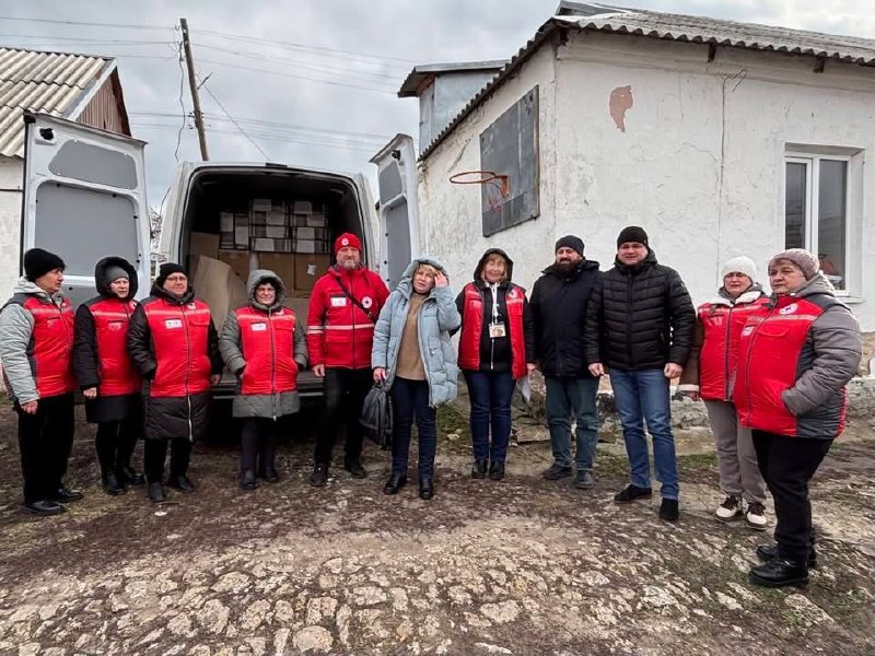 Група людей стоїть на подвір’ї біля відкритого вантажного автомобіля, заповненого коробками. Більшість учасників одягнені в червоні жилети з символікою Червоного Хреста. Поруч розташована одноповерхова будівля з баскетбольним кільцем на стіні.