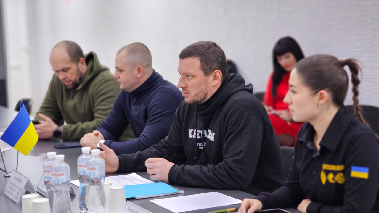 Робоча зустріч у приміщенні з групою людей за столом. На передньому плані чоловік у чорному худі з написом «KHERSON» та жінка у чорній сорочці з нашивкою українського прапора, за ними — ще двоє чоловіків. На столі стоять пляшки з водою, папери та маленький прапорець України.