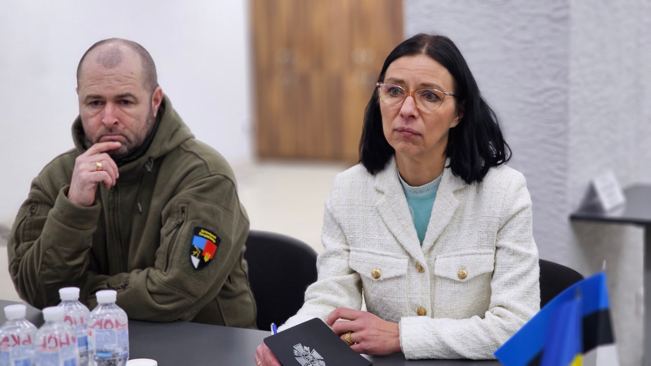 Жінка в окулярах та світлому жакеті сидить за столом поруч із чоловіком у тактичній куртці оливкового кольору. На столі перед ними стоять пляшки з водою та розміщено маленькі прапорці України та Естонії. Жінка тримає в руках блокнот із гербом, обидва учасники зустрічі виглядають зосередженими.