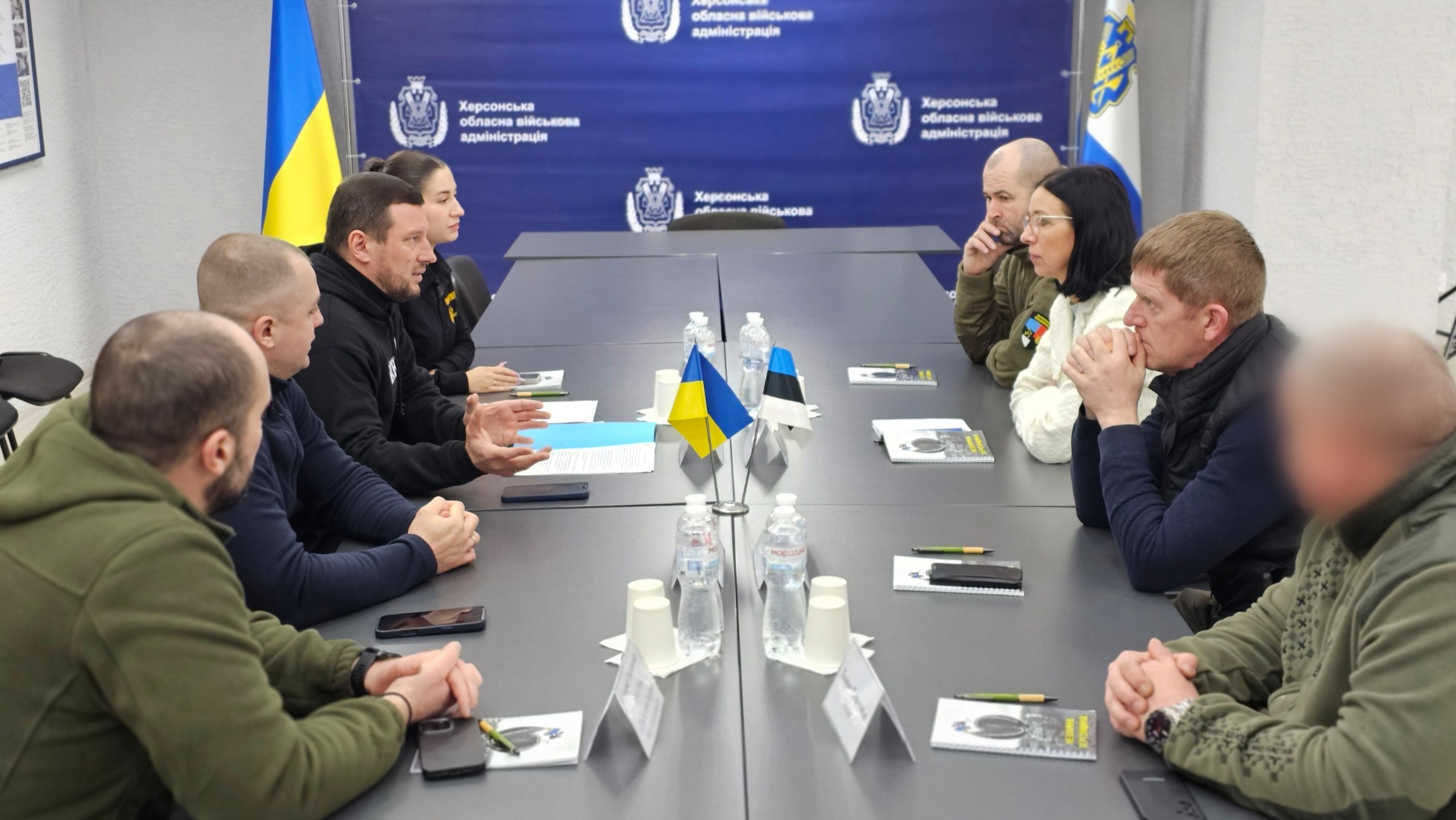 Робоча зустріч двох делегацій за довгим столом у офіційному приміщенні. Учасники сидять один навпроти одного, обговорюючи робочі питання; на столі розміщені маленькі прапорці України та Естонії, блокноти й пляшки з водою. На фоні видно темно-синій банер із гербом та написом «Херсонська обласна військова адміністрація».