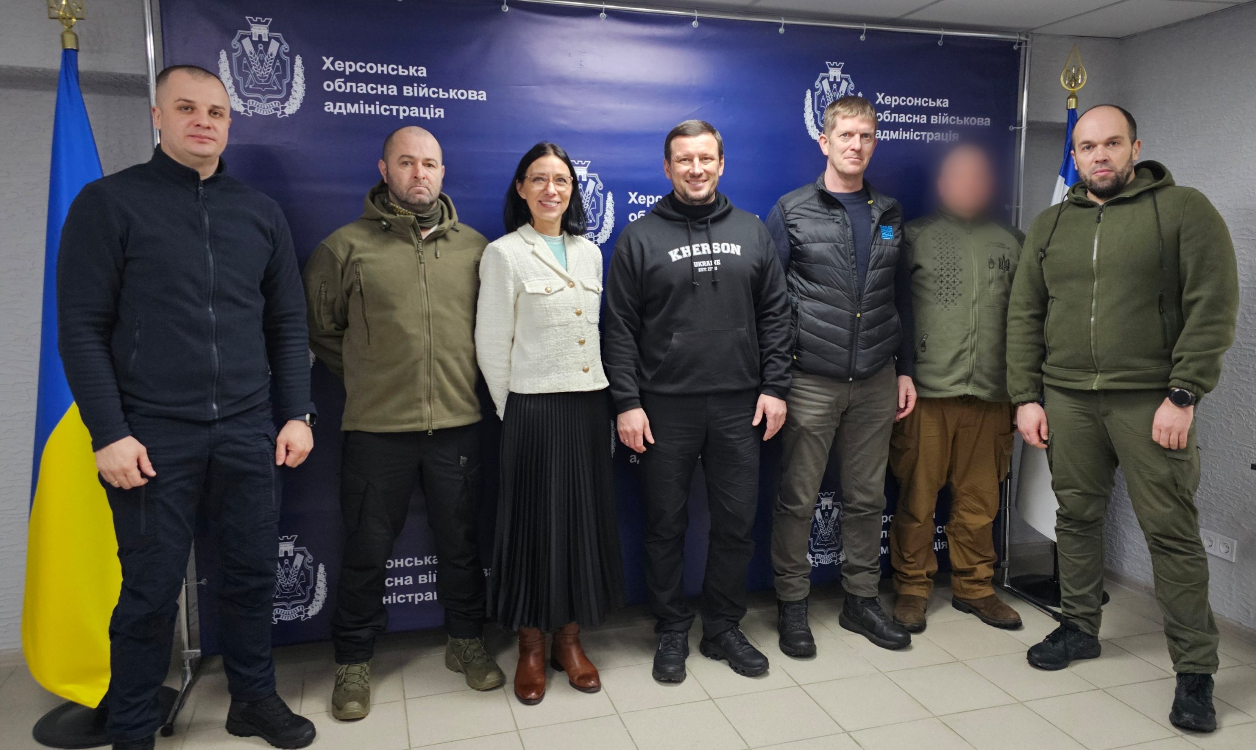 Група людей стоїть у приміщенні на фоні темно-синього банера з логотипом Херсонської обласної військової адміністрації. У центрі стоїть жінка у світлому жакеті та довгій темній спідниці, оточена чоловіками, більшість з яких одягнені у тактичний одяг або худі. Зліва від групи встановлено державний прапор України.