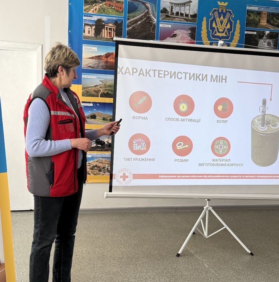 Спікерка в червоному жилеті проводить презентацію на тему «Характеристики мін». На екрані продемонстровано інфографіку з іконками, що описують форму, спосіб активації, колір та інші параметри вибухонебезпечних предметів. На задньому плані розміщено плакат із фотографіями визначних місць Херсонщини.