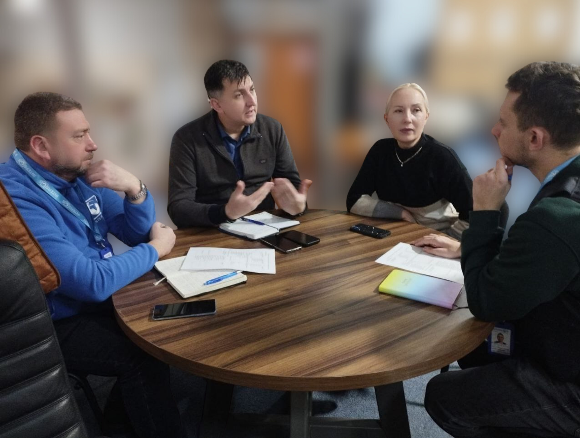 Фотографія робочої наради. Четверо колег (троє чоловіків та одна жінка) сидять за круглим столом з документами. Один із чоловіків активно жестикулює, пояснюючи свою думку.