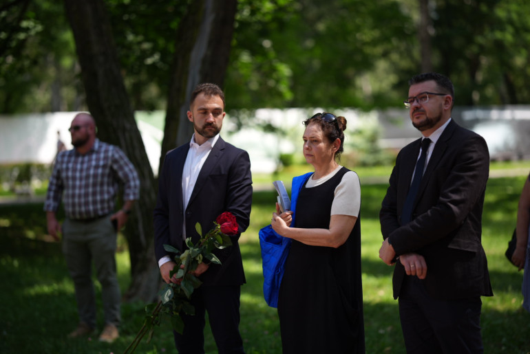 Троє людей у діловому одязі стоять у парку під час офіційного заходу. Чоловік зліва тримає червоні троянди, жінка в центрі тримає синю сумку та папери, а чоловік справа склав руки перед собою. На фоні дерев видно ще одну особу в картатій сорочці.