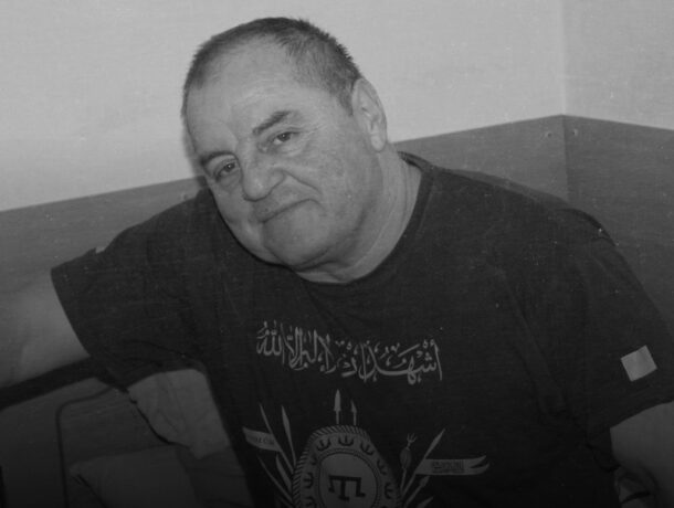 Чорно-білий крупний план обличчя усміхненого чоловіка в темній футболці з арабським написом.