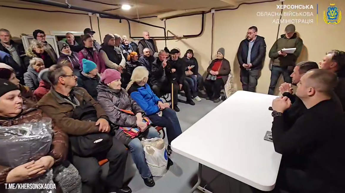 Група з кількох десятків цивільних осіб сидить у підвальному приміщенні або бомбосховищі, ймовірно, під час зустрічі. У правому куті зображення за білим столом сидять чоловіки у цивільному одязі, що спілкуються з присутніми. У верхньому правому куті видно логотип та напис: Херсонська обласна військова адміністрація.