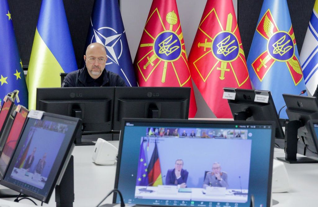 Офіційний представник України сидить за робочим столом перед моніторами під час засідання у форматі відеоконференції. На задньому плані встановлені державний прапор України, прапори Європейського Союзу, НАТО та військові знамена. На передньому плані видно екрани з трансляцією іноземних партнерів.