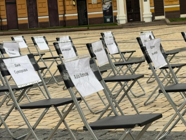 Ряди порожніх розкладних стільців на площі, до спинок яких прикріплені таблички з іменами, такими як: Яна Суворова, Азіз Ахіяров та ін.