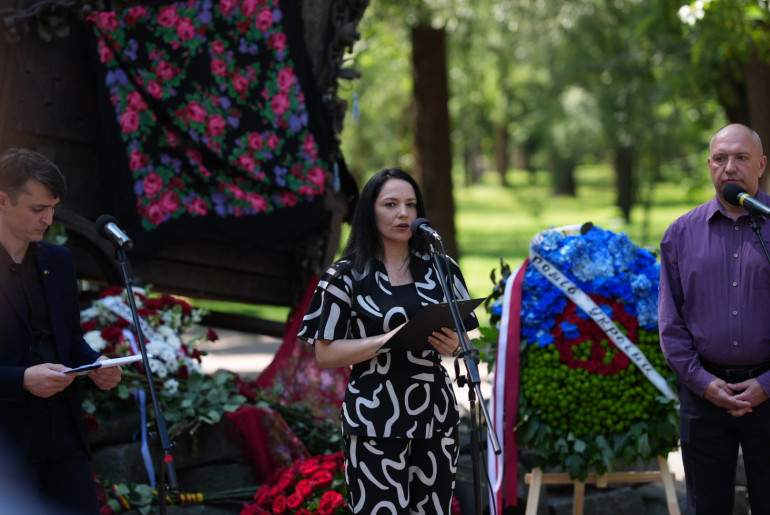 Жінка виступає з промовою біля мікрофонів на меморіальному заході просто неба. Позаду неї стоїть пам'ятник у вигляді залізної кибитки, прикрашений традиційною хусткою, та великий квітковий вінок.