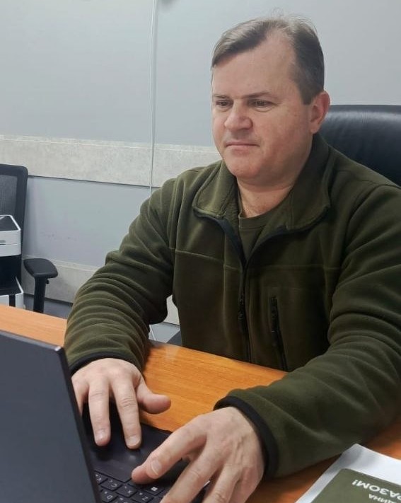 Чоловік у флісовій кофті кольору хакі сидить за робочим столом в офісному кріслі та працює за ноутбуком.