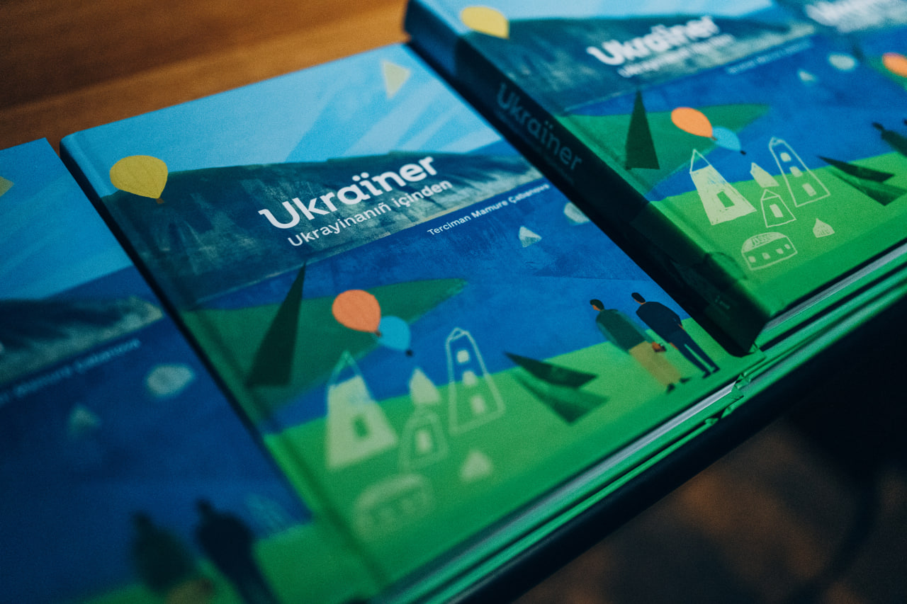 Крупний план декількох примірників книги «Ukraïner» з ілюстрованою обкладинкою в синьо-зелених тонах, що лежать на столі.