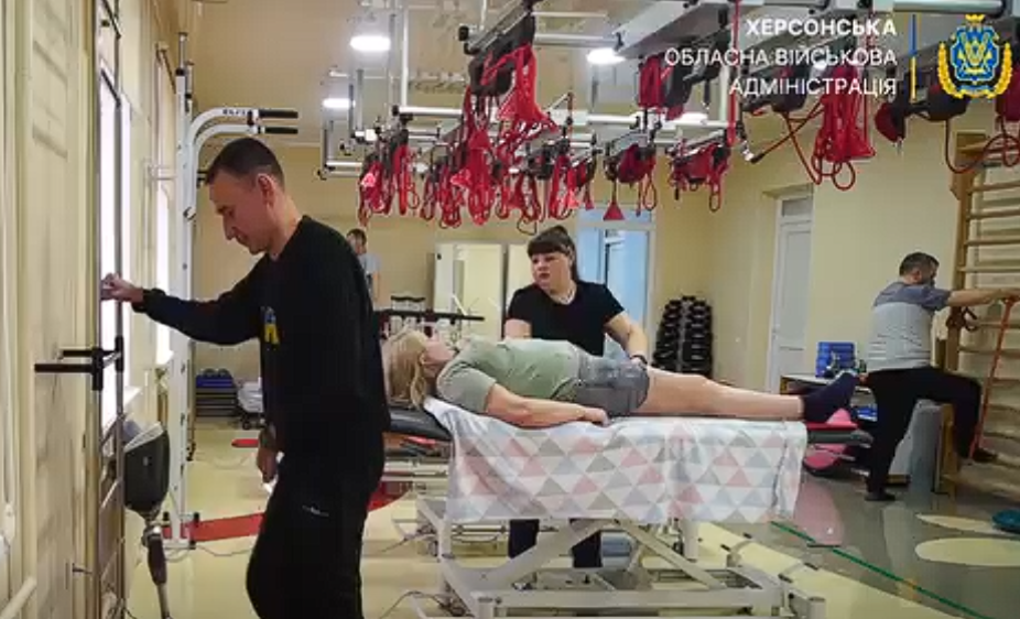 Процес реабілітації у медичному закладі. Фахівці допомагають пацієнтам виконувати вправи у спеціально обладнаному залі з тренажерами та підвісними системами. У кутку стоїть протез ноги. У верхньому правому куті — логотип Херсонської обласної військової адміністрації.